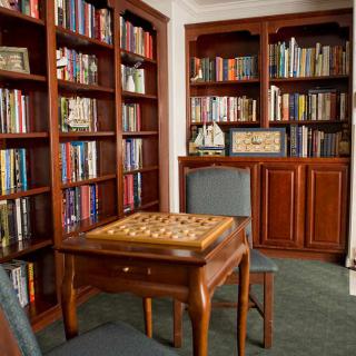A cozy library room featuring wooden bookshelves filled with books and a chessboard on a table surrounded by chairs.