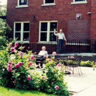Two people enjoying a sunny day in a garden with blooming flowers and outdoor seating in front of a brick building.