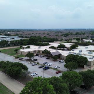 An aerial view of a school surrounded by green trees and a parking lot in a suburban setting.