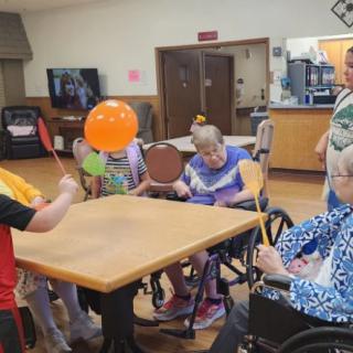 A group of elderly individuals engaging in a balloon activity indoors, seated around a table with some in wheelchairs.