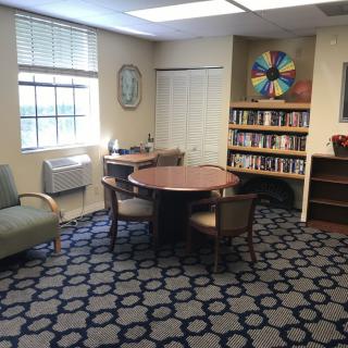 A cozy library corner with a round table, chairs, and bookshelves under natural light from a window.