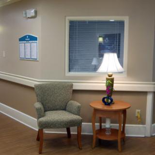 A cozy waiting area featuring a comfortable chair, a small table with a lamp, and soft wall colors.