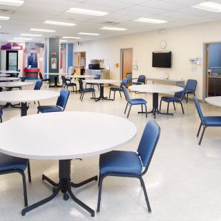 An empty cafeteria with round tables and blue chairs, well-lit by ceiling lights.