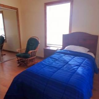 A minimalist bedroom featuring a single bed with a blue blanket, a rocking chair, and wooden flooring.