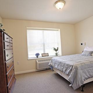 A simple bedroom with a bed, dresser, and window, featuring neutral colors and minimalist decor.