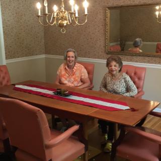 Two individuals seated at a dining table with a chandelier above and a mirror on the wall.