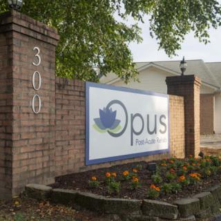 A business sign for Opus displayed on a brick wall near the entrance of a building, surrounded by landscaping.