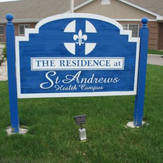 A blue sign for The Residence at St. Andrews Health Campus stands on a grassy patch outside a building.