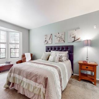 A cozy bedroom with a stylish bed, decorative pillows, and wall art above the headboard. A nightstand with a lamp sits beside the bed, near a large window that brightens the room.