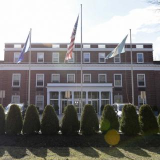 A brick building with multiple flags and neatly trimmed hedges in the foreground.