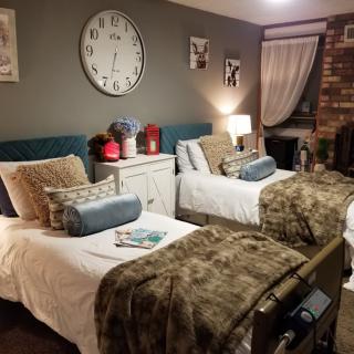 A cozy bedroom with two twin beds, decorated with soft pillows and warm blankets. The room features a large wall clock, a bedside lamp, and a brick accent wall.