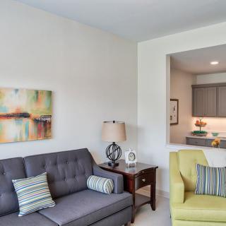 A cozy living room with a gray sofa and yellow armchair, featuring modern artwork and a view into the kitchen.