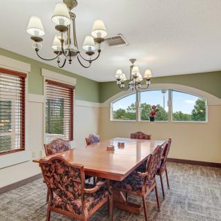 A well-lit dining room with a wooden table and six chairs, featuring chandeliers and large windows.