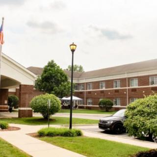 A view of a two-story brick building with a car parked in front, surrounded by neatly trimmed bushes and trees, with flags visible near the entrance.