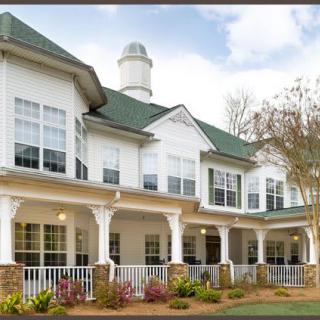 A large two-story residential building with a green roof and white exterior, featuring a spacious porch and well-maintained landscaping.