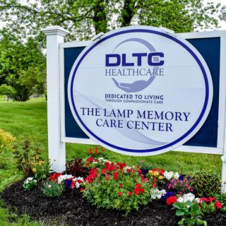 Sign for The Lamp Memory Care Center surrounded by flowers and greenery.