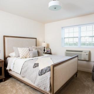 A cozy and well-decorated bedroom featuring a large bed with plush pillows, a side table with a lamp, and a window allowing natural light in.