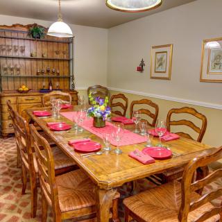 A warmly lit dining room with a large wooden table set for eight, featuring red placemats and a floral centerpiece.