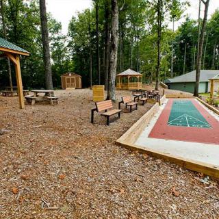 An outdoor recreational area featuring a shuffleboard court surrounded by benches and picnic spots amidst tall trees.