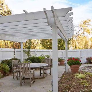 A patio with a white pergola and outdoor wooden seating surrounded by plants.