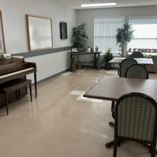 An empty room with a piano, tables, and chairs, featuring large windows and potted plants.