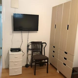 A small bedroom corner with a wall-mounted television, a chair, a tall wardrobe, and a set of drawers.