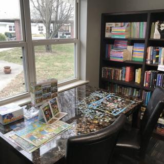 A cozy reading nook with a puzzle in progress on the table, surrounded by bookshelves and natural light from the window.