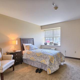 A cozy bedroom with a single bed, nightstand, lamp, and chair, illuminated by natural light from the window.