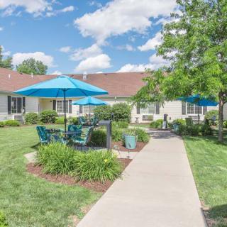 A sunny patio area with tables, chairs, and blue umbrellas surrounded by a well-maintained garden.