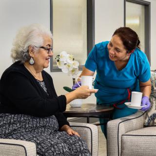A nurse providing care to an elderly woman, serving her a cup of tea in a healthcare facility.