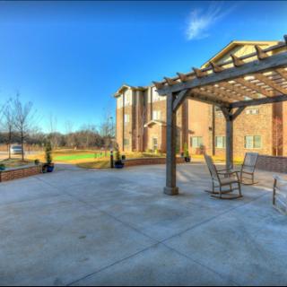A serene outdoor patio area featuring rocking chairs under a pergola, surrounded by lush greenery and a clear blue sky.