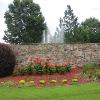A landscaped garden with vibrant flowers in front of a stone wall, featuring a fountain and surrounded by trees.