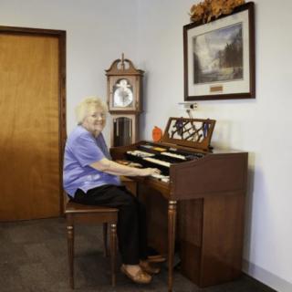 An elderly woman plays the piano in a cozy room, adorned with a clock and artwork on the wall.