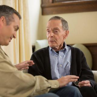 An elderly man and another person engage in a conversation while seated indoors.