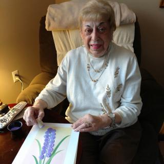 An elderly woman sitting indoors, holding a piece of floral artwork.