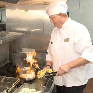 A chef expertly handles a dish on the stove with flames rising in a professional kitchen.