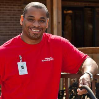 A person smiling outdoors while wearing a red shirt and an ID badge.
