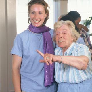 Two people are smiling and interacting indoors, dressed in casual clothing.