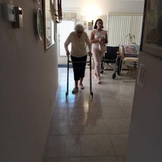 An elderly woman uses a walker while being supported by another woman in a welcoming indoor setting.