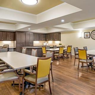 A spacious dining area with multiple tables and chairs, featuring wooden flooring and a kitchen section with cabinets.