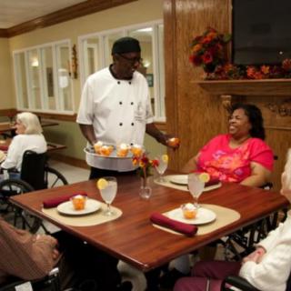 A caregiver serves meals to elderly residents in a nursing home dining area.