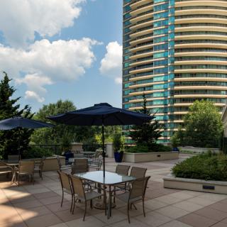 A modern patio with tables and umbrellas next to a high-rise building under a partly cloudy sky.