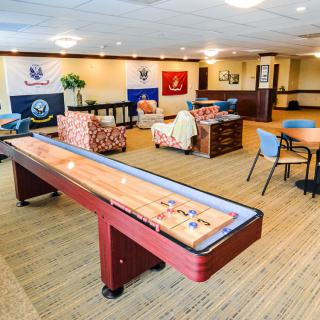 A spacious game room featuring a shuffleboard table, comfortable seating, and various flags on the wall.