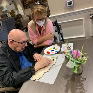 A caregiver assists an elderly man in painting, highlighting the importance of creative activities for mental well-being.
