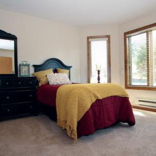 A cozy bedroom with a single bed adorned with burgundy and yellow bedding, featuring a dresser and large windows letting in natural light.