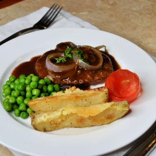 A plated meal featuring steak with gravy and onions, accompanied by peas, tomato, and potato wedges.