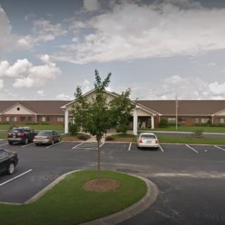 A single-story building with a parking lot in front, featuring several cars and a tree, under a cloudy sky.