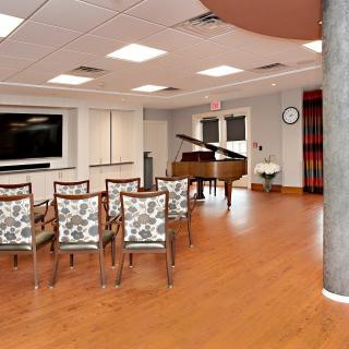 A modern room with chairs arranged facing a grand piano and a large television on the wall.