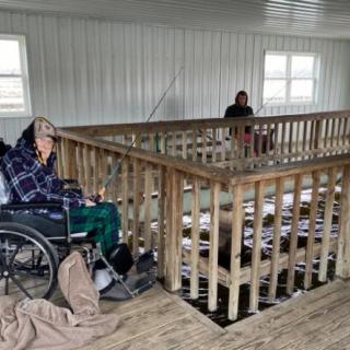 Two individuals, one in a wheelchair and another seated, are indoors with wooden railings.