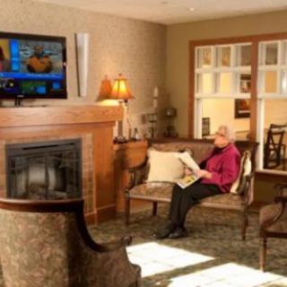 An elderly person sits in a cozy living room reading a newspaper, with a television on the wall and a fireplace nearby.